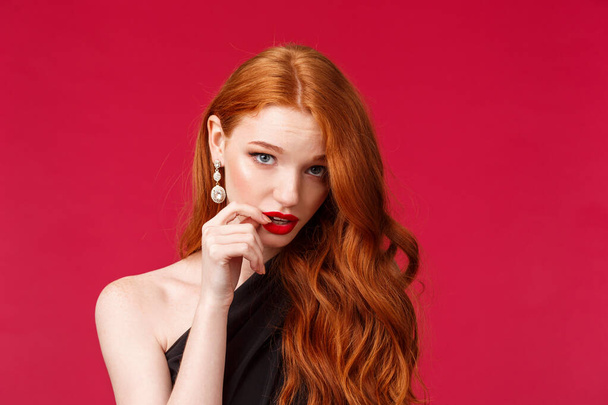 Close-up portrait of silly pretty feminine redhead woman with earrings, red lipstick and black dress, look from under forehead flirty, seducing someone acting clueless and dumb, red background - Photo, Image