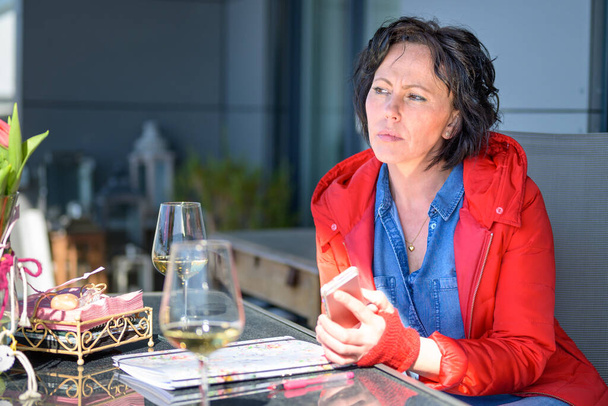 Sorgenvolle trendige Frau in buntem Outfit sitzt vor einem Restaurant und hält ein Glas Weißwein in der Hand und schaut mit nachdenklichem Gesichtsausdruck weg - Foto, Bild