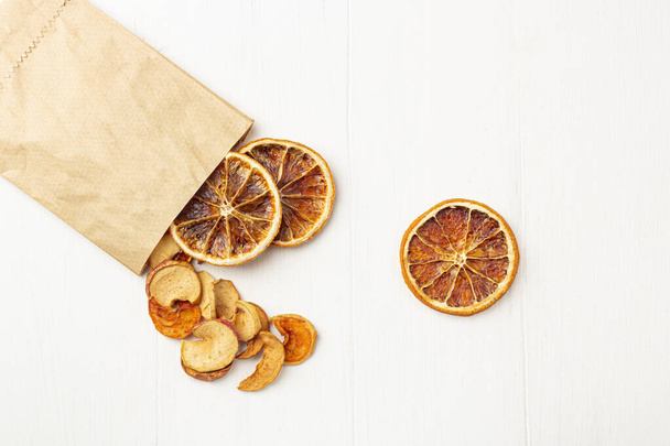 Organic homemade dry fruit chips in a paper eco pack on a white background. Healthy vegan snack of apples and orange. The concept of proper nutrition, organic and vegetarian food. Top view, copy space - 写真・画像