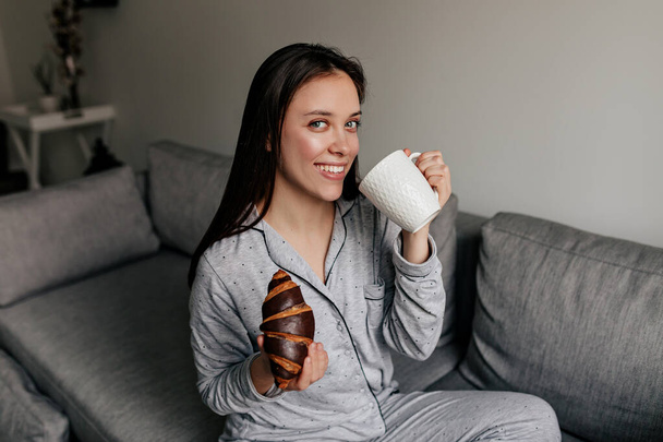 Harika gülüşü olan, pijamalı, kahve içen ve güneşli bir günde evde kruvasan yiyen çekici bir kadın..  - Fotoğraf, Görsel