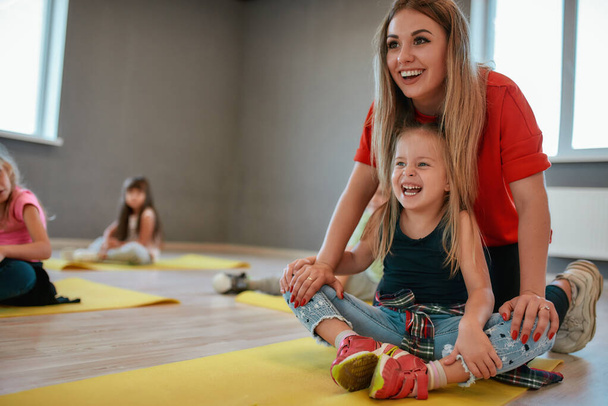 Training a little child. Portrait of a cheerful female trainer helping cute little girl to do butterfly exercise. Group of children sitting on the floor and stretching muscles - Photo, Image