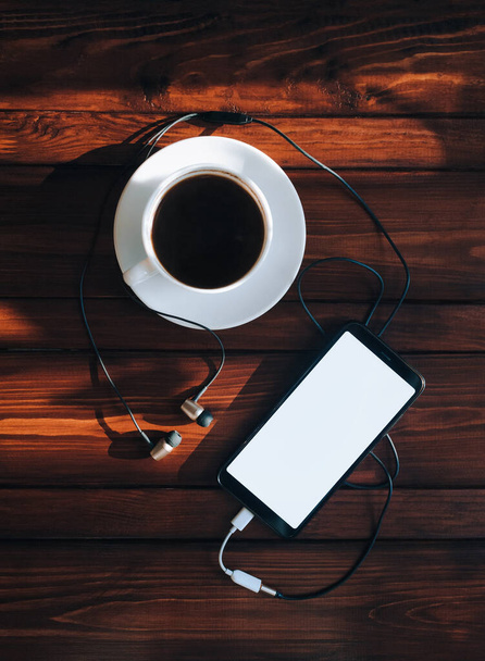 Black smartphone with white screen, dark headphone and cup of coffee lies on a brown wooden background mahogany with spots of sunlight. Top view, copy space, mockup. Morning breakfast, music concept. - Photo, image