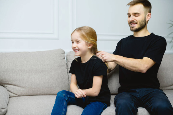 Young father at home with his cute little daughter making her a hairstyle. Similar casual outfit - black t-shirt and jeans. - Zdjęcie, obraz
