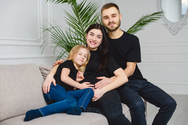 赤ちゃんを期待して黒い服の幸せな家族。リビングルームのソファに座っている間、若い家族、父、妊婦の母親と彼らの小さな娘は抱っこしています。カメラを見ている家族. - 写真・画像