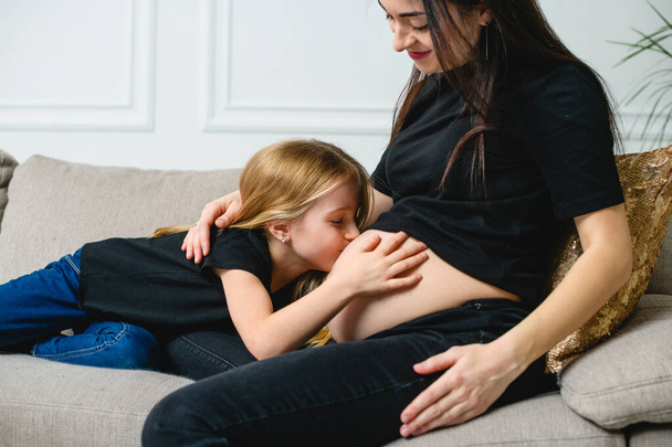 Girl kiss mother pregnant tummy at couch. Sweet pregnant mom with daughter on sofa. - Photo, Image