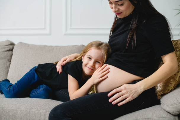 Mutter und Mädchen sitzen am Sofa und berühren Bauch, glückliche Familie von zwei werdenden Babys. Ähnliches lässiges Outfit - schwarzes T-Shirt und Jeans. - Foto, Bild