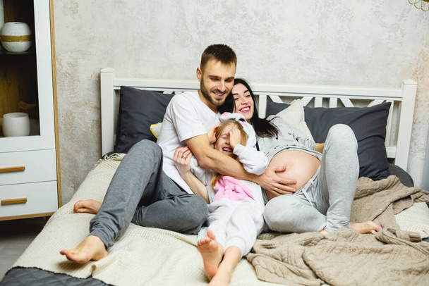 Szczęśliwy ojciec, córka i ciężarna matka przytulają się w łóżku w domu pijamas w sypialni. - Zdjęcie, obraz