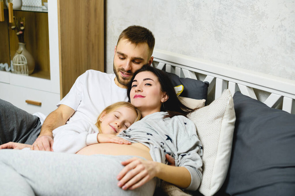 Portret pięknej rodziny oczekującej dziecka. Szczęśliwy ojciec, córka i ciężarna matka przytulają się w łóżku w domu pijamas w sypialni. - Zdjęcie, obraz