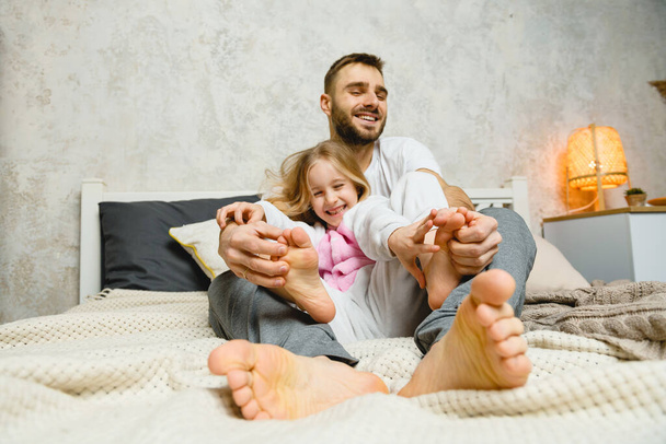 Young father tickles smiling daugther in bed. Dad and daughter tickle each other's feet. - Photo, Image
