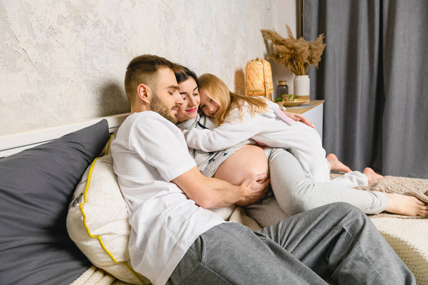 ベッドで幸せな家族。パジャマを着た幸せな父親、娘、妊娠中の母親。夫は妊娠中の妻を抱きしめる。娘は母親を抱きしめる. - 写真・画像