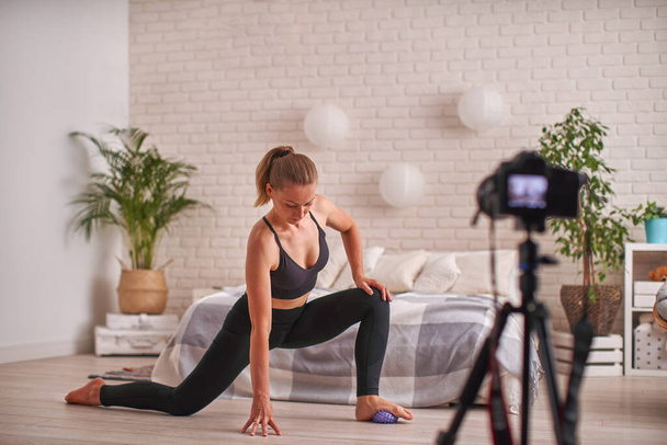 Athletic woman blogger in sportswear shoots video on camera as she does exercises at home in living room. Sport and recreation concept. Healthy lifestyle. Fitness vlogger recording live tutorial video - 写真・画像