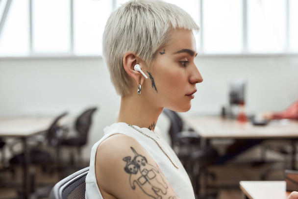 Side view of young and cute blonde tattooed business woman with short haircut wearing wireless earphones while working in the modern office. Video, skype call - Фото, зображення