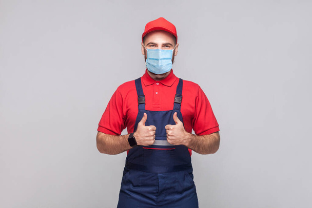 Work are done! Portrait of young man with surgical medical mask in blue overall, red t-shirt , cap, standing and showing thumps up and looking at camera. Grey background, indoor studio shot isolated. - Foto, imagen