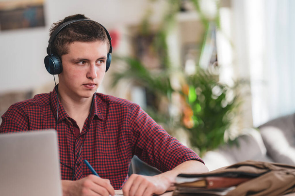 Brooding active teen guy with headphones listens to lecture, does in notebook, using laptop and makes notes in Notepad. concept of online learning. Social distance, self-isolation. Distance learning. - Foto, Bild