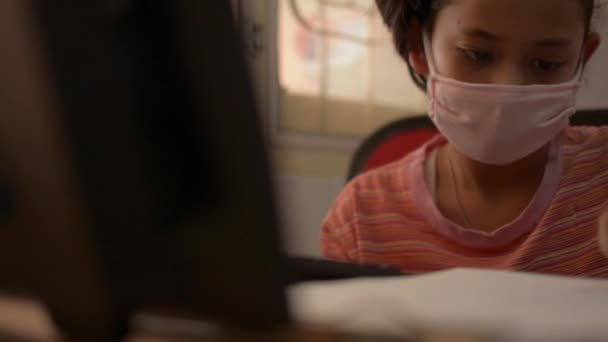 Χαριτωμένο κορίτσι φορώντας προστατευτική μάσκα και γράφοντας στο γραφείο με τον υπολογιστή στο σπίτι κατά τη διάρκεια της επιδημίας COVID-19 κατάσταση. Μελέτη στο σπίτι έννοια. - Πλάνα, βίντεο