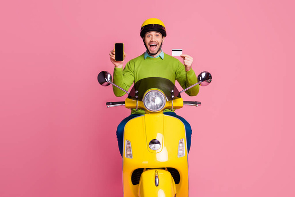 Portrait of his he nice attractive glad cheerful cheery guy riding moped holding in hands cell bank card fast transfer rent isolated over pink pastel color background - Foto, Imagem