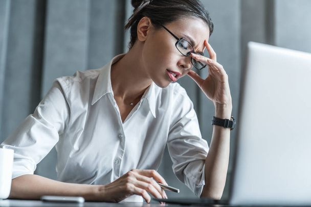 Stressed young female office worker sitting at desk holding head because of pain in office - Foto, Bild
