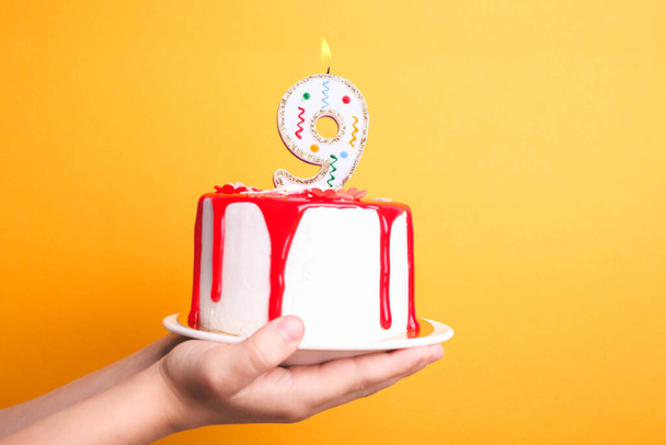 Weiße Sahnetorte mit rotem Zuckerguss und einer Kerze in den Händen der Kinder auf orangefarbenem Hintergrund. Geburtstagskonzept. - Foto, Bild