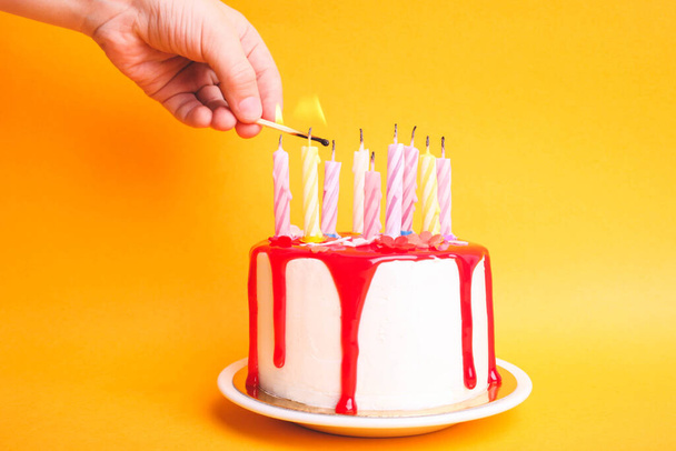 La mano da bambini accende candele su una torta bianca con ciliegina rossa su uno sfondo arancione. Concetto compleanno
. - Foto, immagini