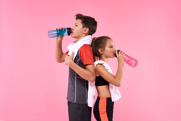 Every Sip Counts. Two teenagers boy and girl posing with towels and bottles of water after workout isolated on pink background. Fitness, sport, training, active lifestyle concept - Foto, Bild