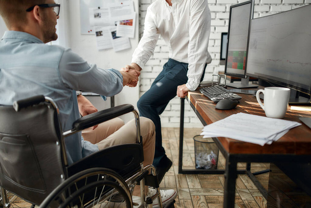 Těší mě. Mladý kancelářský pracovník na invalidním vozíku potřásá rukou svému kolegovi, zatímco pracuje společně v kreativní kanceláři. Koncept zdravotního postižení - Fotografie, Obrázek