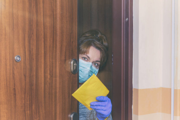 Γυναίκα με προστατευτική μάσκα προσώπου και γάντια που ανοίγουν την πόρτα και λαμβάνουν ταχυδρομική επιστολή, επιδημία του ιού του κόκορα covid-19. Αρχική καραντίνα και αυτο-απομόνωση έννοια - Φωτογραφία, εικόνα