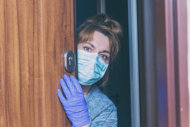 Korumalı yüz maskesi takan ve eldivenli bir kadın Corona virüsü salgını yüzünden evinin kapılarını açıyor. Ev karantinası ve kendini izole etme kavramı - Fotoğraf, Görsel