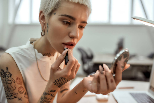 Cute lady at work. Portrait of young and stylish blonde tattooed business woman applying lipstick while sitting in the modern coworking space - Фото, изображение
