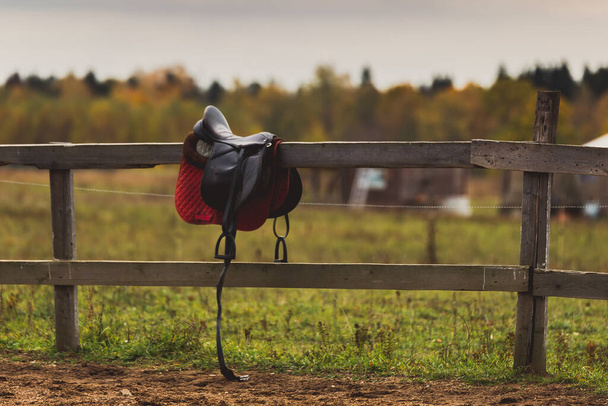 A saddle hangs on a wooden fence on a ranch against the background of a field - 写真・画像