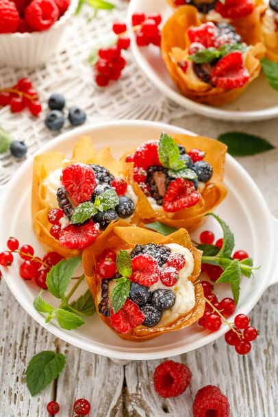 Филловые фруктовые чашки со взбитыми сливками с добавлением свежих ягод, посыпанных сахарной пудрой на белую тарелку. Вкусный десерт из фило - Фото, изображение