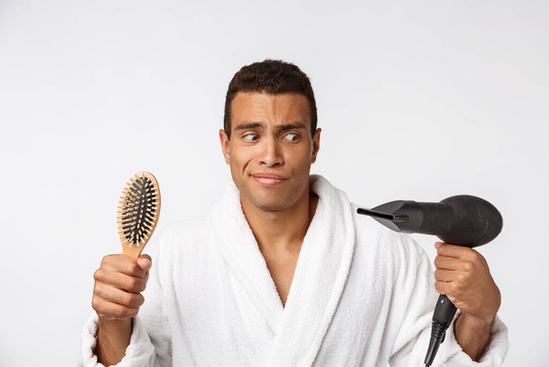 Portrait of mature funny beautiful tan-skinned african guy with curly hair in casual white bathrobe having problems with combing hair and hair dryer - Photo, Image