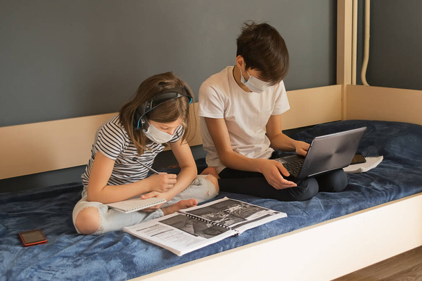 Παιδιά αγόρι και ένα κορίτσι με ιατρική μάσκα κάνουν μαθήματα μέσω φορητού υπολογιστή. Ηλεκτρονική εκπαίδευση κατά τη διάρκεια επιδημίας πανδημίας. - Φωτογραφία, εικόνα