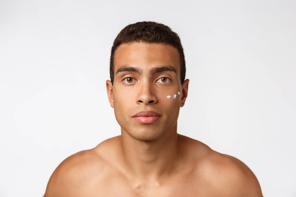Foto de hombre afroamericano sin camisa sonriendo y aplicando crema facial aislada sobre fondo blanco
 - Foto, imagen
