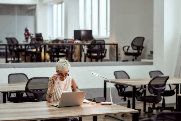 Business lady at work. Young focused blonde tattooed woman in eyewear working with laptop while sitting alone in the modern office - Photo, Image