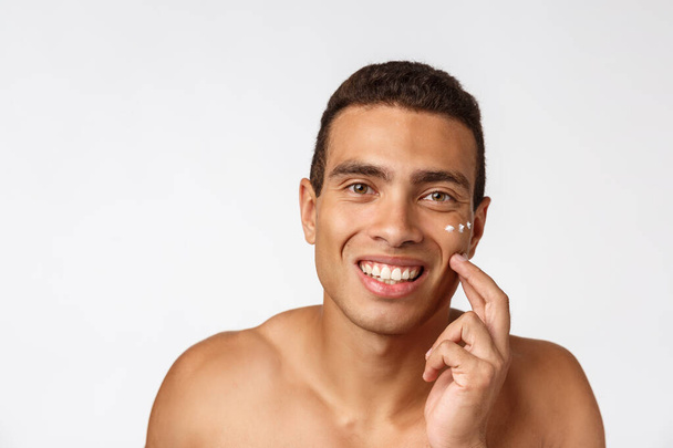 Foto de homem americano africano sem camisa sorrindo e aplicando creme facial isolado sobre fundo branco
 - Foto, Imagem