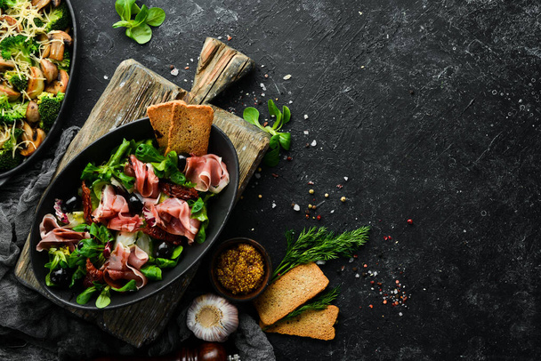 Prosciutto salad with olives in a black bowl. Spanish cuisine. Top view. Free space for your text. Rustic style. - Foto, immagini