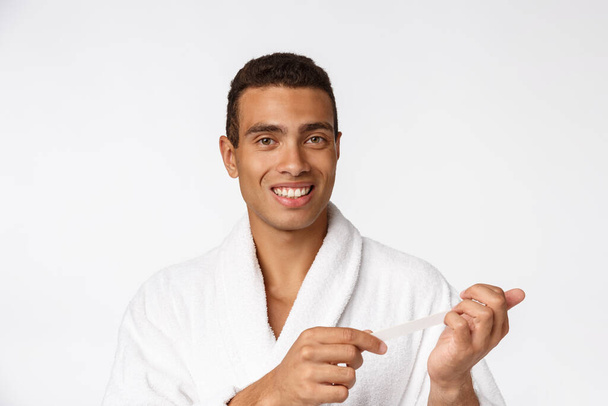 Cooler gutaussehender Mann im Bademantel, der Maniküre für sich selbst macht, mit Nagelfeile isoliert über weißem Hintergrund. - Foto, Bild