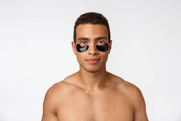 Atractivo joven africano con parches negros bajo los ojos cuidando su piel facial mirando directamente a la cámara sobre fondo gris
 - Foto, Imagen