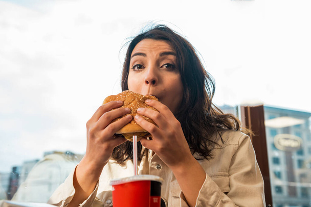 Menina bonito bonito bonito bonita em roupas da moda goza de um hambúrguer fast food grande e bebida enquanto sentado em um café
. - Foto, Imagem