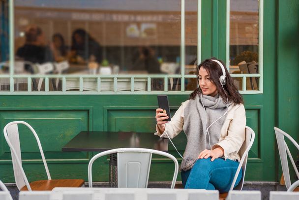 Beautiful young girl in fashionable clothes listen music with headphones and takes selfie while sitting at a cafe outside.Lifestyle concept - Foto, immagini