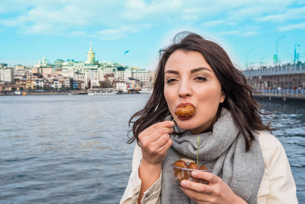 Moda kıyafetleri içinde güzel bir kadın, Türkiye 'nin İstanbul kentindeki Galata Kulesi, Galata Köprüsü ve Boğaz manzaralı geleneksel Türk tatlısını yiyor - Fotoğraf, Görsel