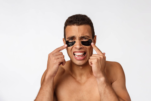 Attraktiver afrikanischer junger Mann mit schwarzen Flecken unter den Augen, der seine Gesichtshaut pflegt und direkt in die Kamera auf grauem Hintergrund blickt - Foto, Bild