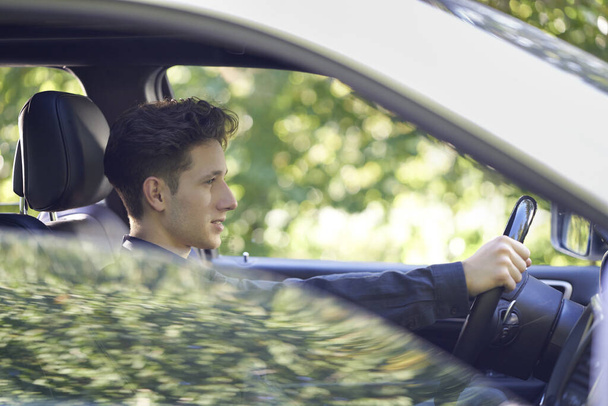 Perfil de um jovem motorista bonito sentado em seu carro com as mãos ao volante, a janela lateral meio aberta e com reflexos de luz
                      - Foto, Imagem
