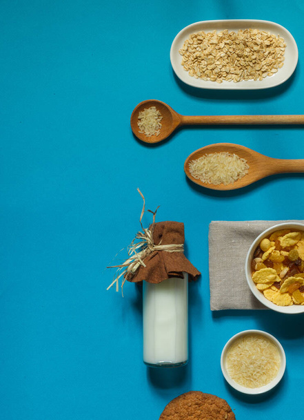 platte ingrediënten voor een snel en gezond ontbijt - rijstmelk in een fles, cornflakes en gedroogd fruit op een helderblauwe achtergrond met horizontale ruimte - Foto, afbeelding