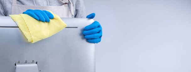 La joven ama de llaves en delantal está limpiando la superficie de la computadora plateada con guantes azules, trapo amarillo húmedo, primer plano, espacio para copiar, concepto de diseño en blanco
. - Foto, Imagen