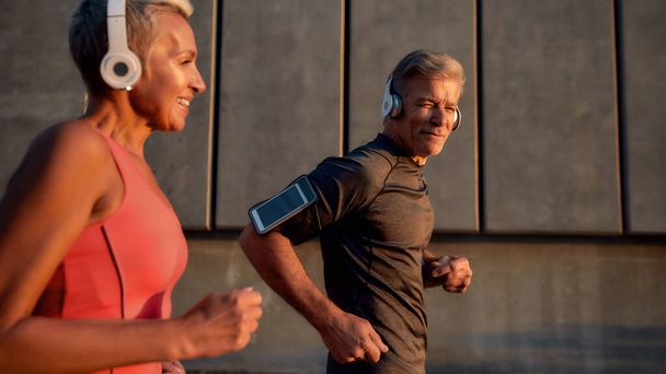 Sport making us happier. Positive and active middle-aged couple in headphones running through the city street in the morning - Photo, Image