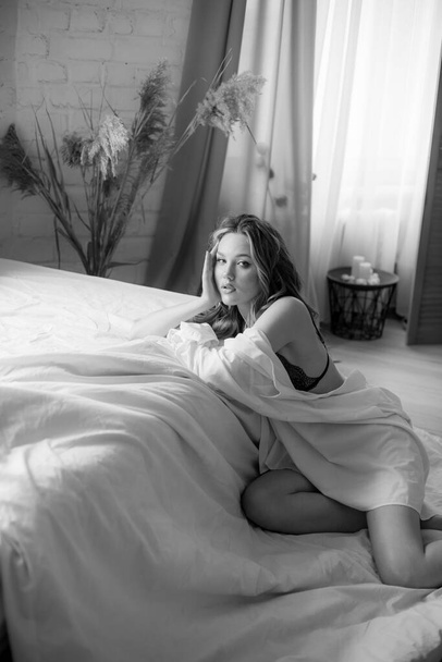 Lovely young girl with long curly blond hair in the morning in the bedroom, in a beautiful lace negligee and white shirt. - Fotografie, Obrázek