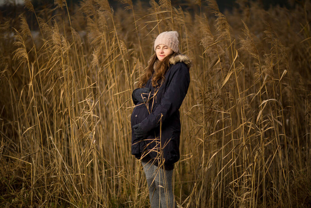 Mujer embarazada en un bosque o hierba en otoño o invierno. Foto del embarazo de una mujer de pie en una hierba alta. usar sombrero chaqueta frío si
 - Foto, imagen