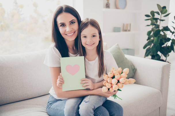 Photo of pretty little girl young charming mommy sitting cozy sofa holding hands 8 march present postcard tulips flowers from daddy affection emotions casual clothing home indoors - Фото, зображення
