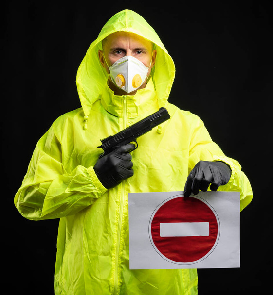  Covid-19 Coronavirus-Infektionskonzept. Mann in Atemschutzmaske und Handschuhen mit Pistole und Stoppschild vor schwarzem Hintergrund - Foto, Bild